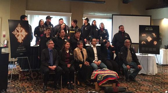 Group shot of several people sitting on chairs as well as standing up at the Feathers of Hope: Justice & Juries report release event. The people include youth and elders. On either side of them is an enlarged poster of the artwork from the Justice and Juries report. Each poster is propped up on an easel. In front of the group of people is a Mother Drum wrapped in a blanket.