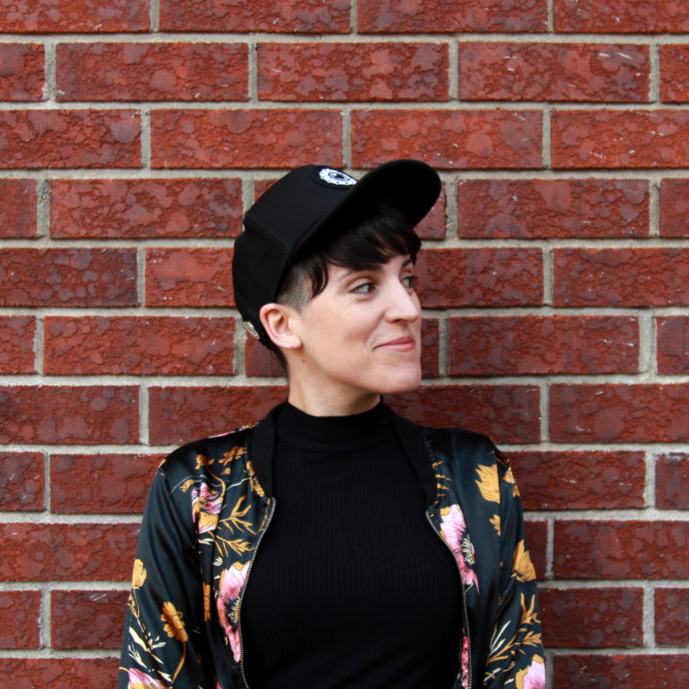 Photo of Guadalupe Pérez Pita, a Latinx person with short hair. Guadalupe is wearing a black baseball cap and a black floral jacket. They are standing in front of a brick wall.