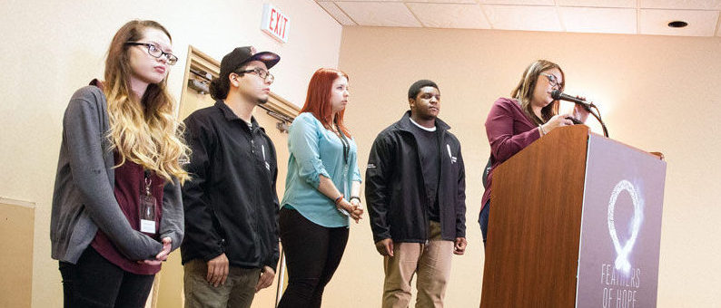 Four Youth Amplifiers and one Youth Advisor who worked on the Justice and Juries report. The youth on the far right hand side is at a podium speaking into a microphone while the other four youth stand behind the podium.