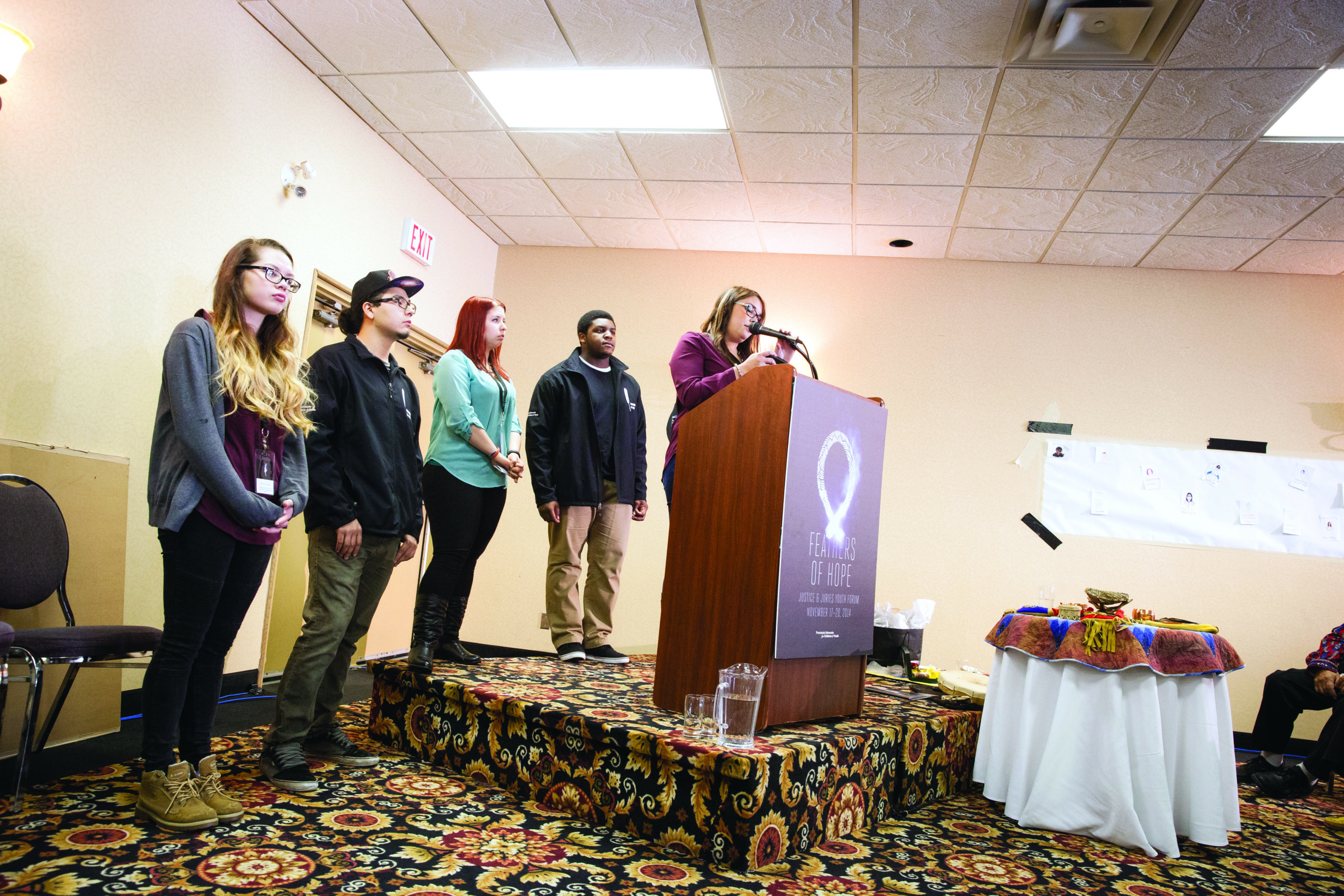 Four Youth Amplifiers and one Youth Advisor who worked on the Justice and Juries report. The youth on the far right hand side is at a podium speaking into a microphone while the other four youth stand behind the podium.