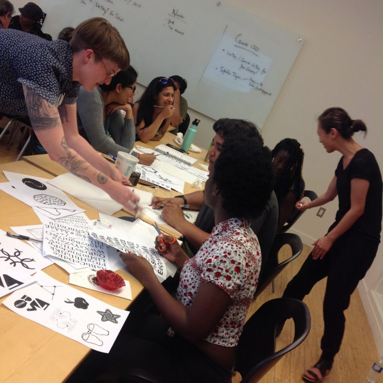 Photo: Several people sitting around wood tables, sketching on pieces of paper and discussing what they are drawing. Two facilitators are standing to the left and right, leaning over the tables to talk to participants. On the tables are printouts of icons and typefaces.
