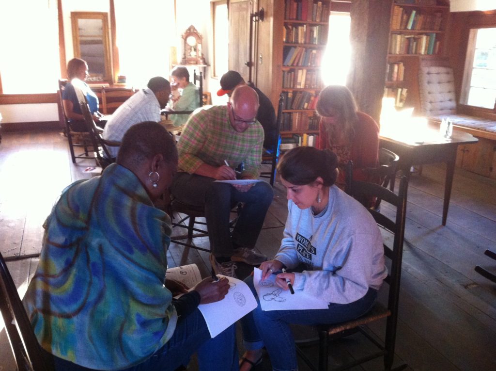 A photo of two people sitting on wooden chairs, facing each other as they make sketches on sheets of paper resting on their laps. The person to the left of the scene is drawing with their back turned to the camera, as the person on the right is leaning toward them, attentively looking at what they draw. More people appear on the background of the photo, pairs of individuals who are also drawing and exchanging ideas about their sketches.