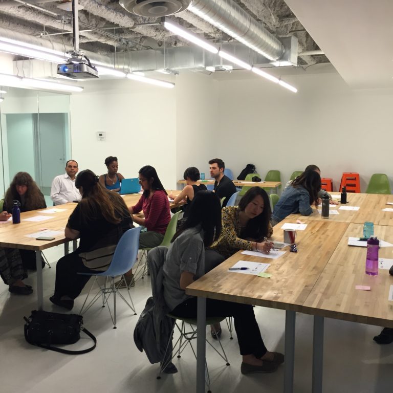 Photo: Several people sitting around wood tables, sketching on pieces of paper and discussing what they are drawing.