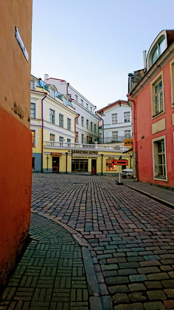This photograph depicts a street view of an old European city. The buildings are stacked, different sized, and go from white to warm tones, the streets are a deeper multi colour cobblestone. The sky is light blue.