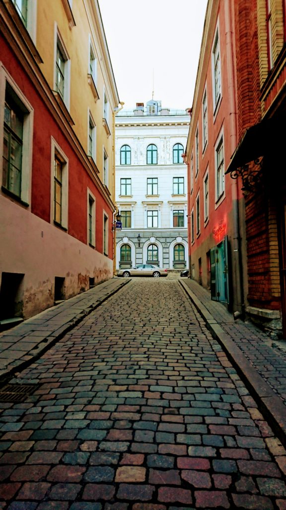 Photo: A European, multi-coloured, cobblestone, alleyway, lined with multi coloured buildings with white window frames. There is a car at the end of the alley and white building adding to the windows. The sky above is a light gray.