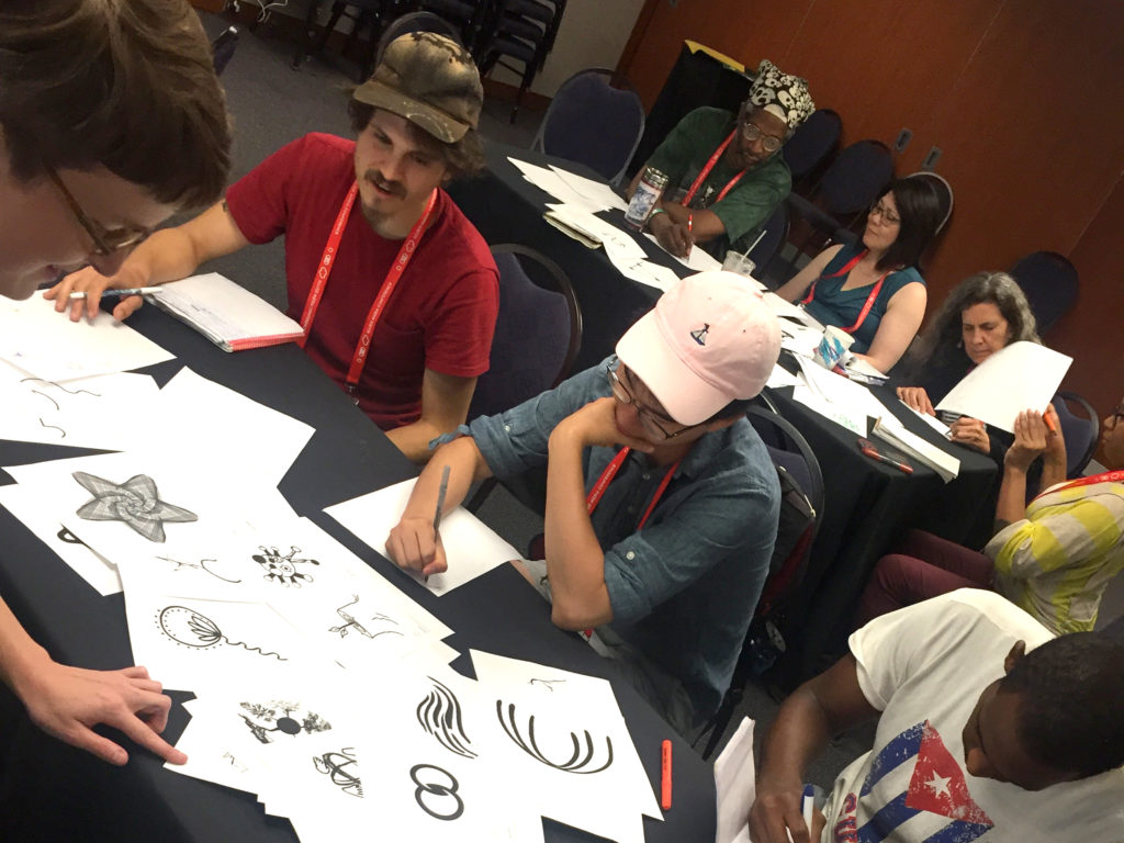 Several people sit at tables, sketching on pieces of paper. In front of them are printouts of icson from the Vision Archive representing concepts like empathy, unity, and more. Some icons are geometric and digital looking, while others have a more hand drawn aesthetic.