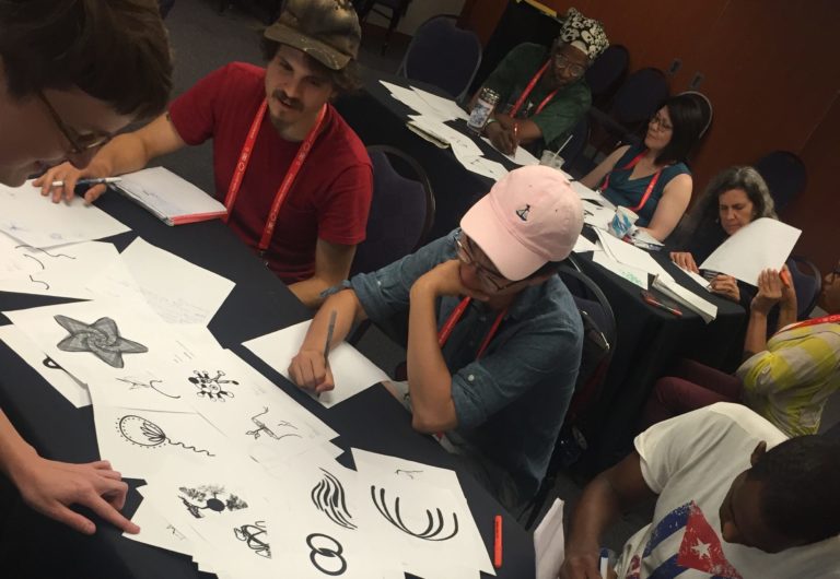 Several people sit at tables, sketching on pieces of paper. In front of them are printouts of icson from the Vision Archive representing concepts like empathy, unity, and more. Some icons are geometric and digital looking, while others have a more hand drawn aesthetic.
