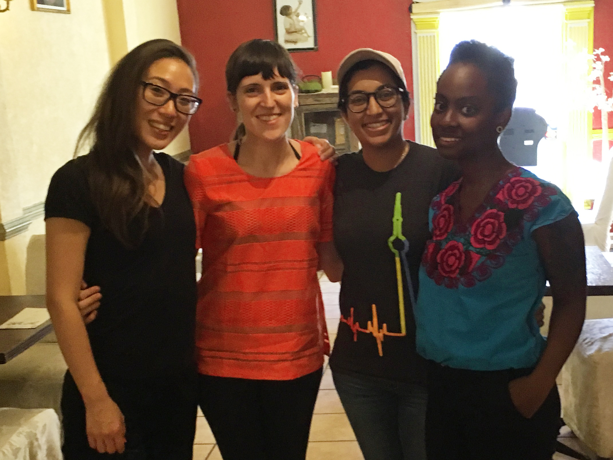 Photo: four women of colour pose arm-in-arm, smiling and looking at the camera.
