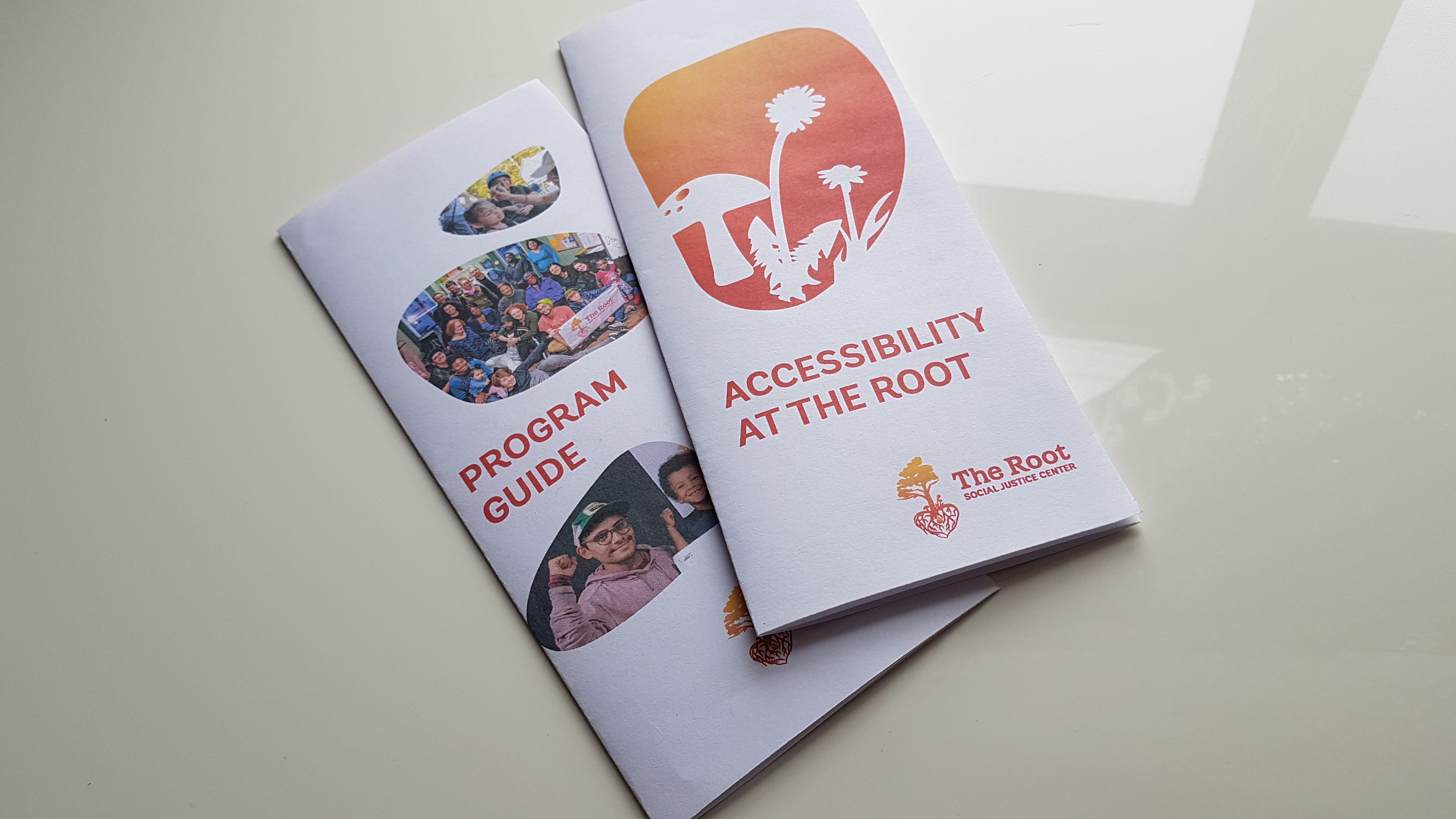 Photo: Two folded brochures sit on a white surface, one stacked on the other. The top brochure reads "Accessibility at The Root" and has silhouettes of a toadstool and dandelions against a gradient gold to red background, inside a rock-shaped frame. In the bottom right hand corner is The Root logo. The one below it reads "Program Guide" and has 3 photos of various people, mainly people of colour, inside of rock shaped frames.