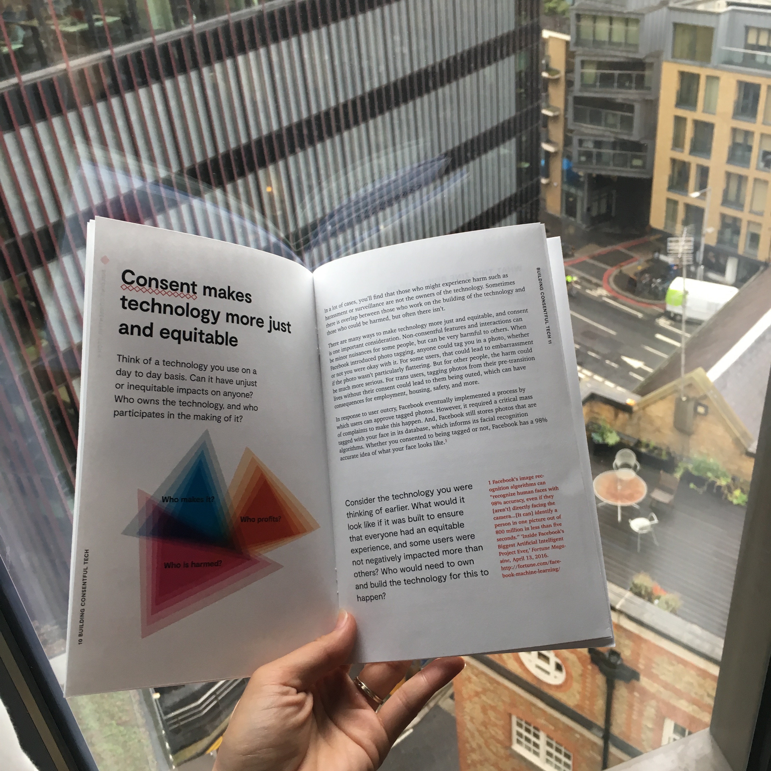 Photo: Una’s left hand holds open the Building Consentful Tech zine. The spread that is visible reads “Consent makes technology more just and equitable” and there is a graphic showing three intersecting triangles with the labels “Who makes it?” “Who profits?” and “Who is harmed?” Behind the zine is a window overlooking a streetscape in London.