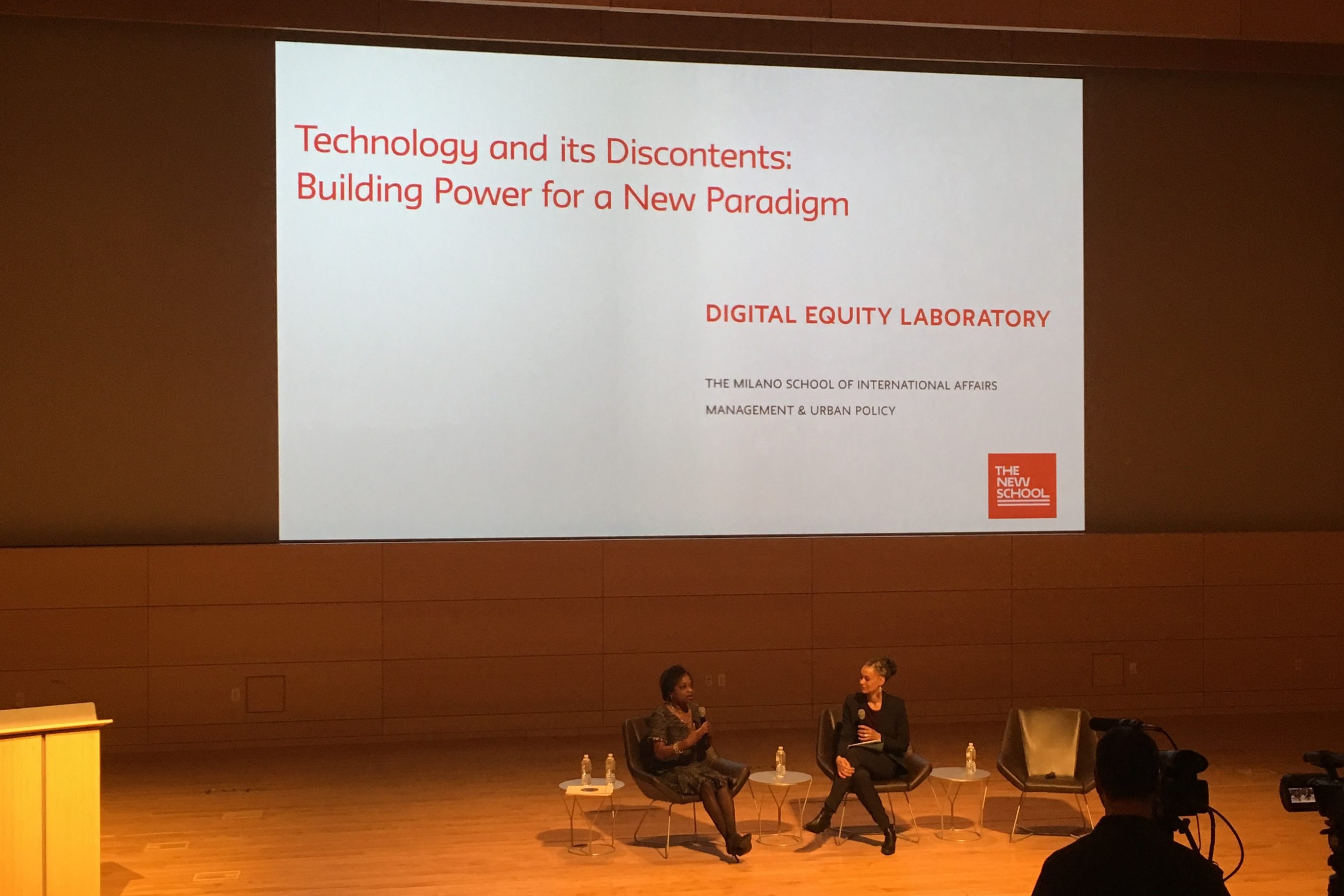 Two African American women are seated and in conversation on a stage. Behind them is a large white screen with the title of the event, "Technology and its Discontents: Building Power for a New Paradigm" in orange.
