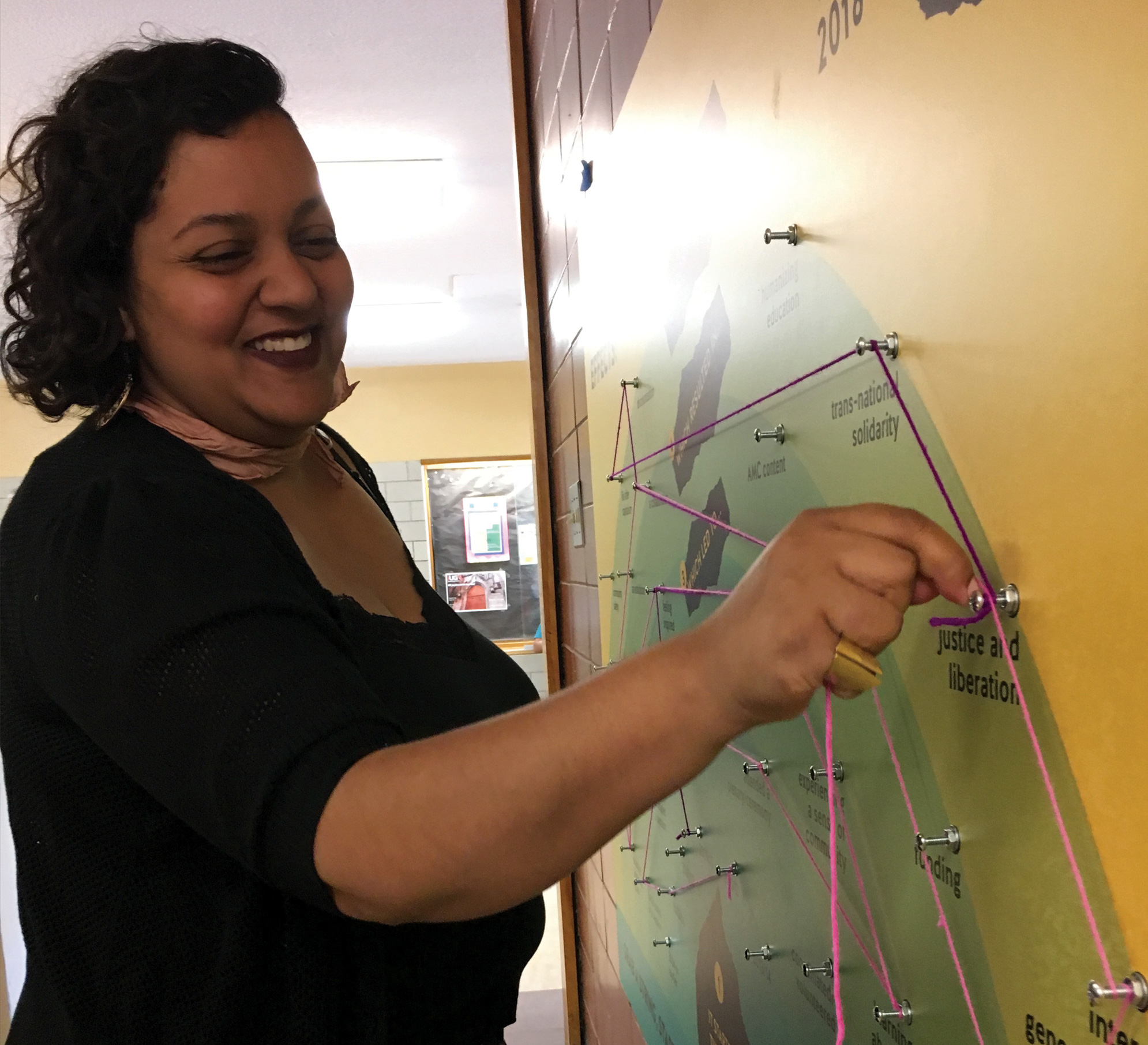 Photo of an AMC attendee standing before the Ripple Mapping Tool as they map their story onto the poster. The person is smiling as they twist a piece of pink string around a nail labelled "justice and liberation."