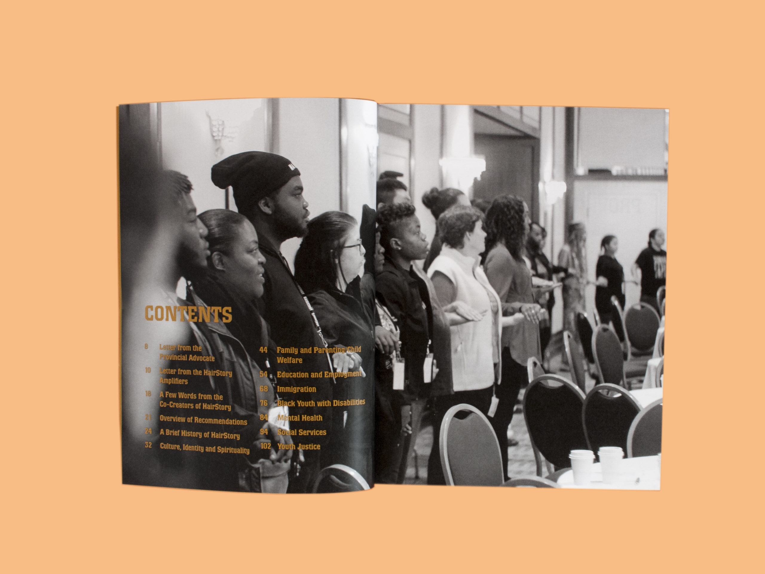 Photo of the contents spread from HairStory—Rooted. Behind the table of contents is a large photograph of many people standing in a circle, holding hands. Most of the people are Black youth, and they stand alongside elders and allies.