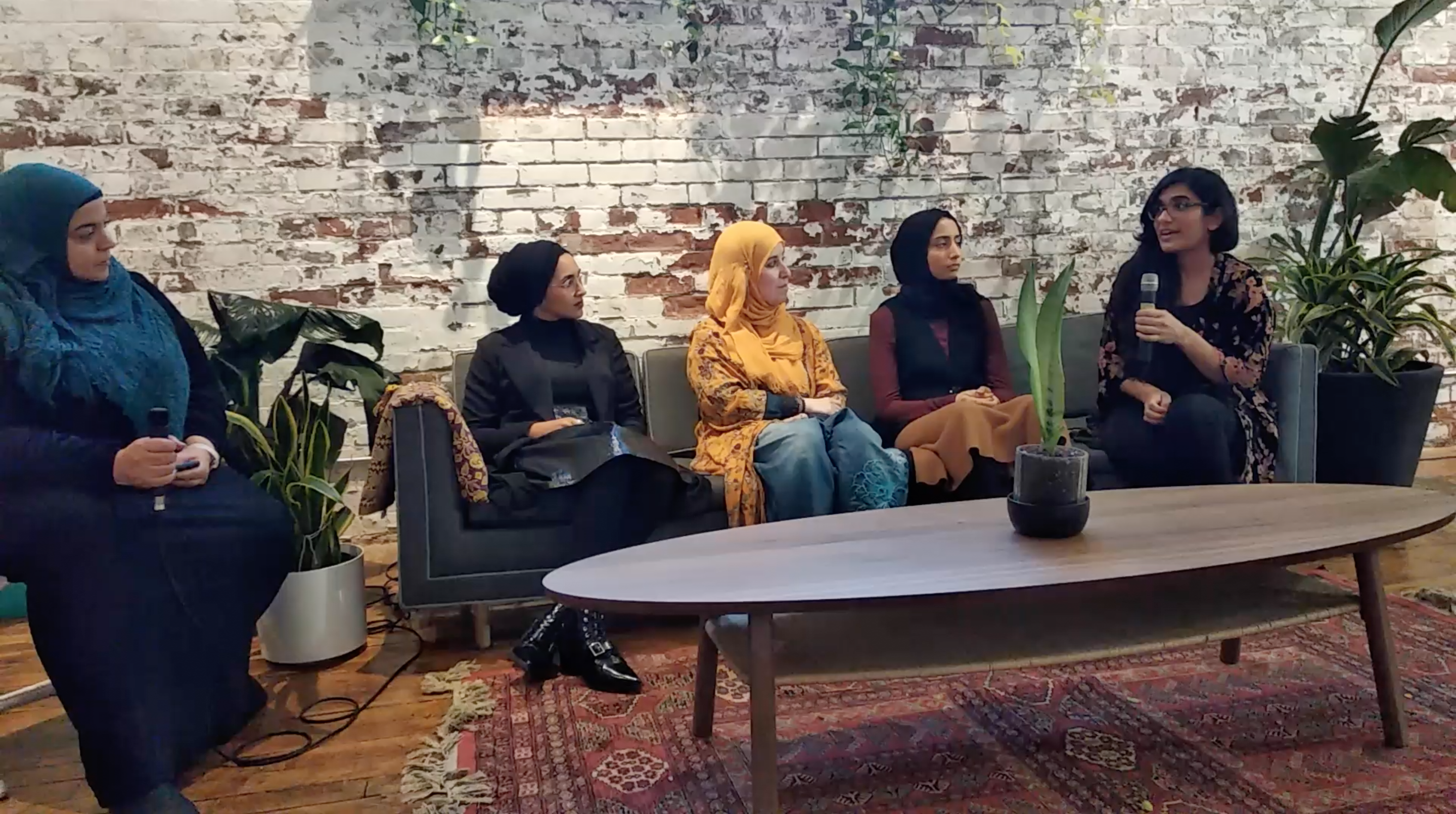 A photograph of 5 people- a moderator and four panelits. The moderator sits on a chair to the left of the photo and four panelists sit on a couch to the right side of the image. The space has a living room feel with a washed brick wall backdrop, there are plants in the space, a warm rug, and a coffee table and plants. 