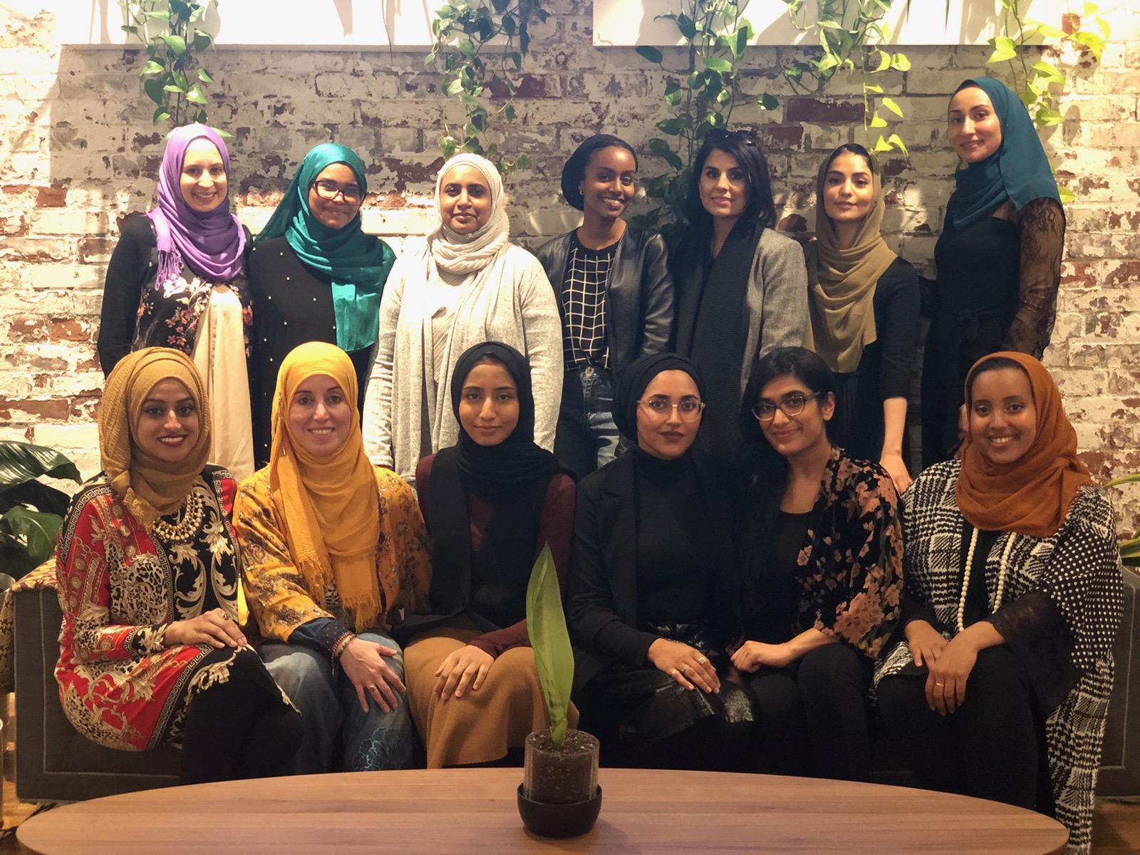 A photograph of women. 7 standing at the back and 6 sitting on a sofa in the middle ground, there is a coffee table with a plant on it in the foreground. The backdrop is a whitewashed brick wall with windows and hanging plants.