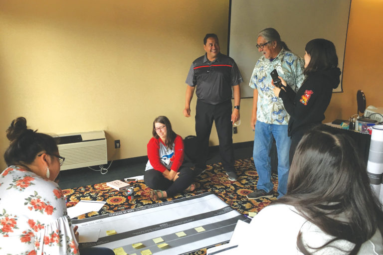 Photo of an Indigenous Elder surrounded by an adult person and four young people, who all listen attentively and with joyful expressions as the Elder speaks. At their feet stands a large sheet of paper placed on the floor, over which is visible a storyline chart with some posts-its on it.
