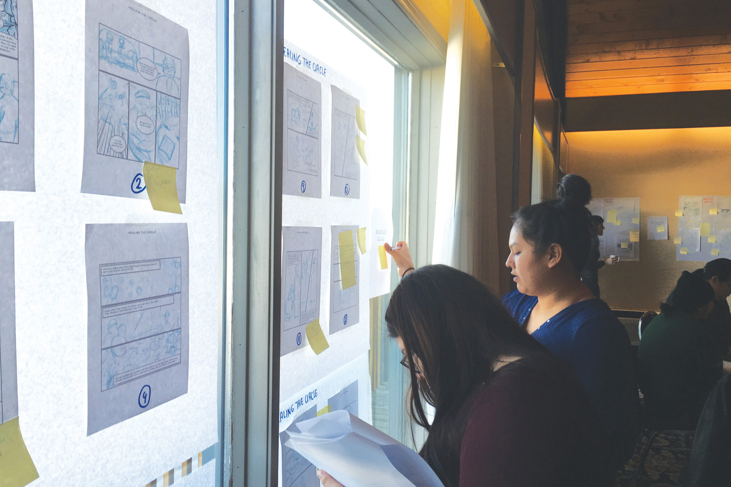 Photo of two young advisors standing in front of a window, to which are taped several graphic novel storyboards. The advisors are giving feedback by placing post-its on individual panels.