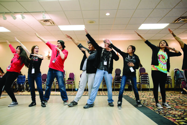 Photo of seven young people standing in a line, each raising their right arm towards the sky. While partaking in this group activity, some of them are looking serious and determined, while some of them are laughing.