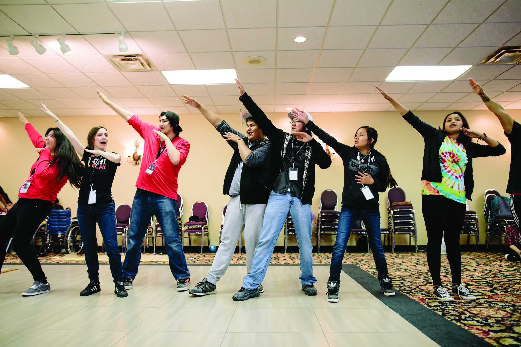 Photo of seven young people standing in a line, each raising their right arm towards the sky. While partaking in this group activity, some of them are looking serious and determined, while some of them are laughing.