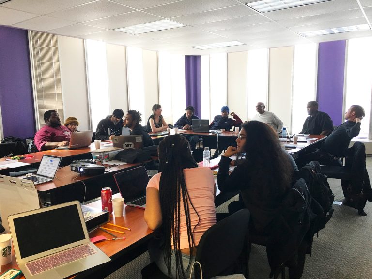 Photo of a group of multigenerational and multiracial people sitting at tables arranged in a circle. The people are huddled in small groups, talking and writing.