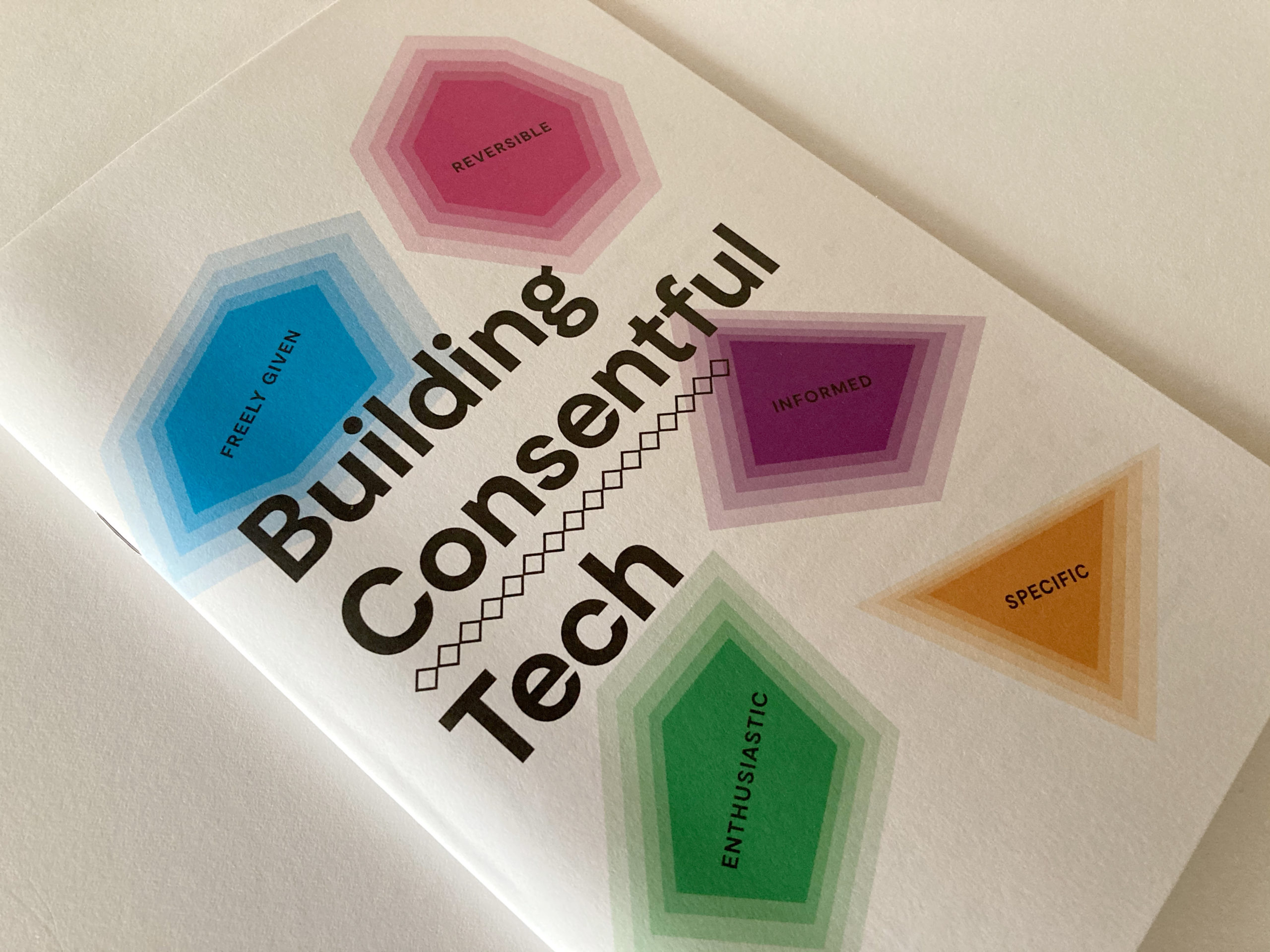 A photograph of the Building Consentful Tech zine on a table. The title is surrounded by gem shapes of different colors, with the words "Freely Given, Reversible, Informed, Enthusiastic, and Specific" inside of them.