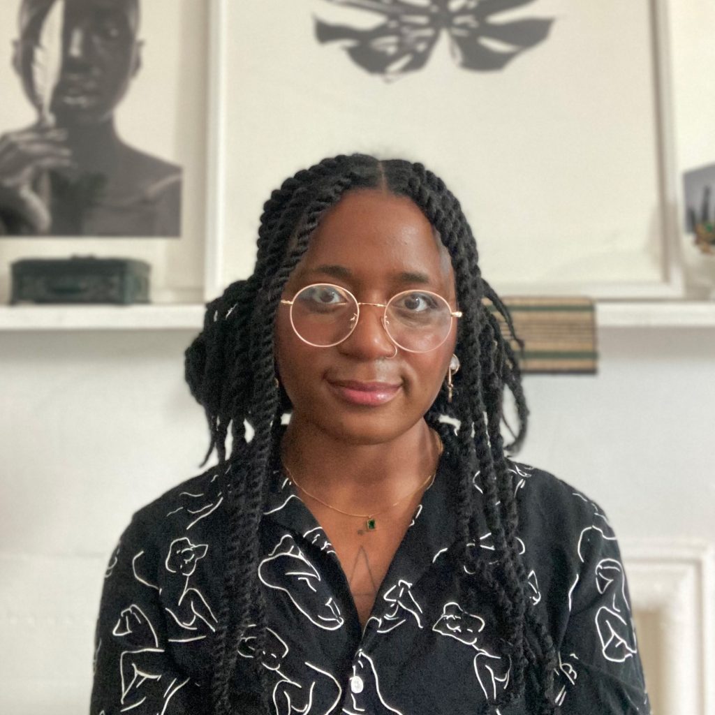 Photograph of Adriana Warner, a young Black woman wearing pink glasses and smiling. Adriana is facing forward, the image is cropped from the chest up. She wears a black and white patterned shirt, hair styled in Senegalese twists. Behind her, the background is primarily white and out of focus. 2 framed prints sit on a shelf, one of a black woman holding a white feather, the other of a monstera leaf.