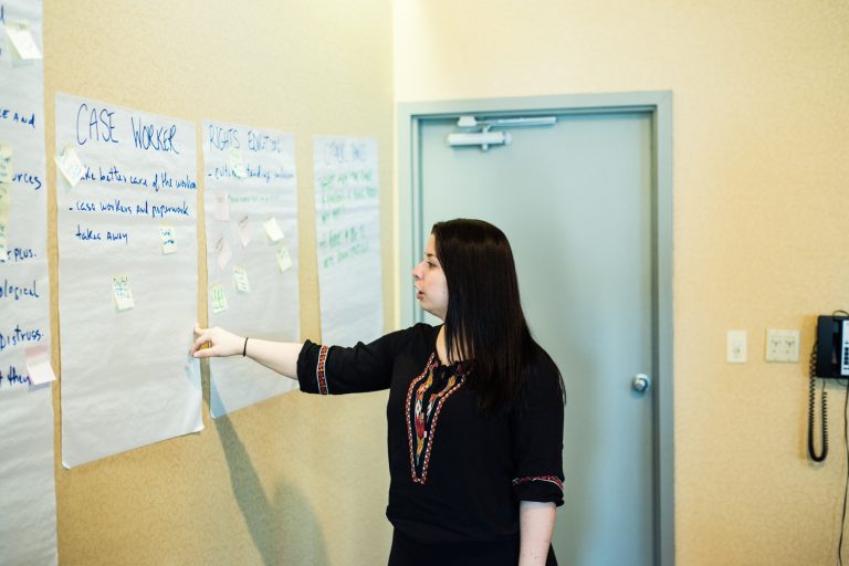 A young Indigenous woman with shoulder length brown hair and a nose piercing stands in front of four sheets of chart paper with sticky notes on them. She is pointing at and talking about one sheet with the title "Case Worker."