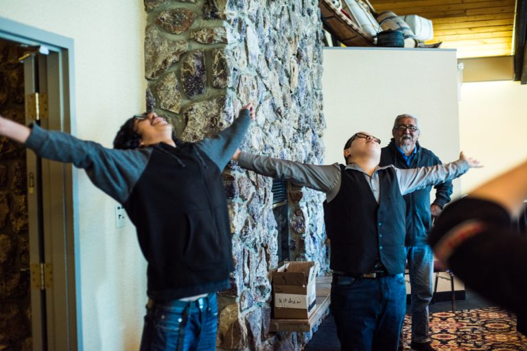 Photo of two young Indigenous men standing with their arms outstretched and faces pointed up at the ceiling. One is smiling, and the other looks peaceful. An elder stands behind them.