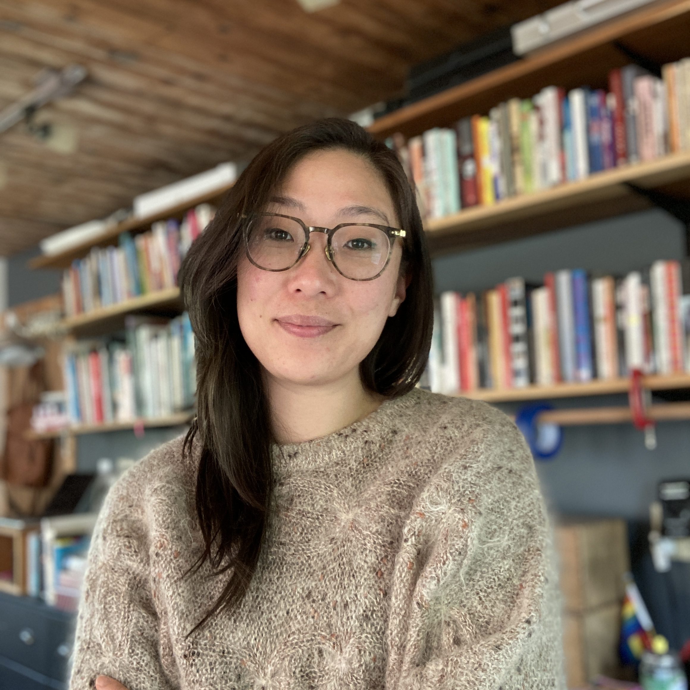Photo of Una Lee, a Korean woman with long hair and glasses. She is wearing a fuzzy beige sweater and standing in front of a bookshelf.