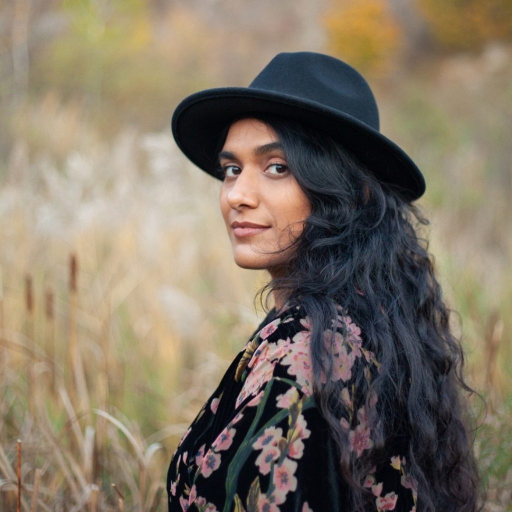 A portrait of Zahra Agjee amidst tall reed, cattail, and grass. She is in the foreground of the photo with only her upper body in the frame. Her body faces the left of the frame while face and gaze are toward the camera. She wears a black felt hat and a floral pink, green, and black shirt.