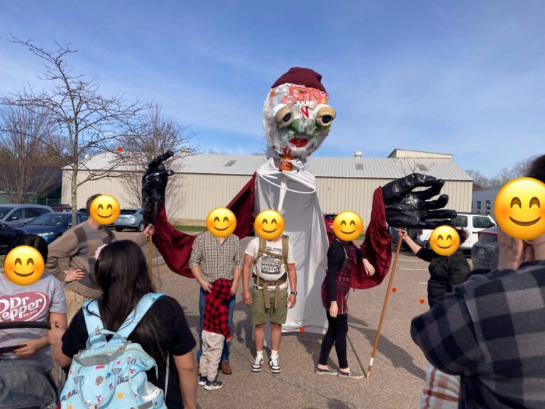A group of people, with faces obscured by emojis, standing with a dairy worker puppet. The puppet is partially finished and has a soulful face made of flattened milk jugs, is wearing a red beanie, and is wearing black gloves.